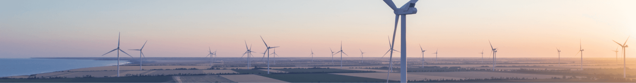 Een windmolenpark met meerdere windturbines op een open veld bij zonsondergang, met uitzicht op de zee aan de horizon.